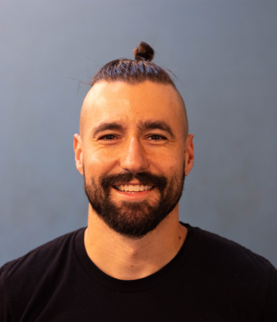 A man with a beard and mustache smiles at the camera. He has dark hair styled in a topknot and shaved sides, wearing a black t-shirt—ready for a session at an Austin Climbing Gym, against a plain blue background.
