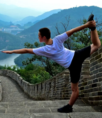 A person in a white shirt and black shorts practices a yoga pose on the Great Wall of China, surrounded by lush green hills and misty mountains in the background.