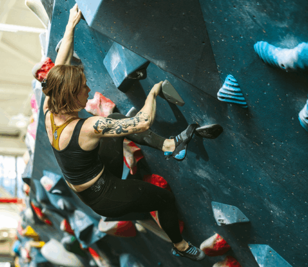 A person with tattoos climbs an indoor bouldering wall, gripping large blue and white holds while wearing climbing shoes and athletic clothing. The wall is steep, ideal for personal coaching sessions in all markets.