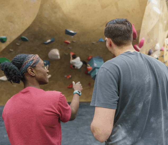 Two people standing indoors, facing a climbing wall with colorful holds. One points toward the wall, seemingly offering Personal Coaching as they discuss climbing strategies. Both appear engaged and focused on the challenging route ahead.