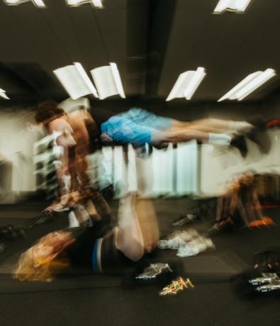 Two people in a gym perform an acro yoga pose; one lies on the ground lifting the other into the air with their legs. The motion blur gives a dynamic, energetic feel to the scene. Gym equipment is visible in the background.