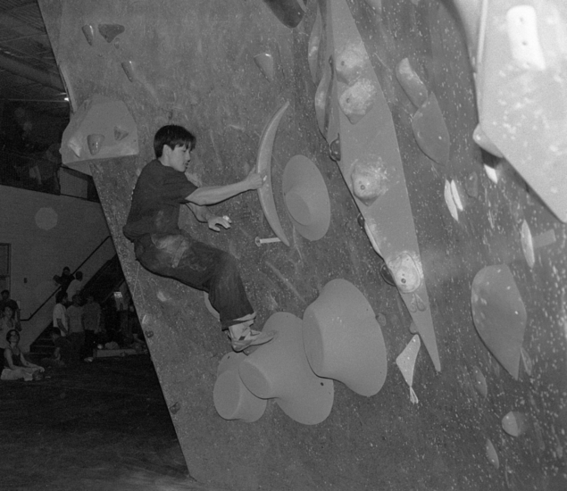 A person is climbing an indoor bouldering wall using large holds, while others, possibly from Boston youth teams, watch in the background. The scene appears to be in a gym or climbing facility. The photo is in black and white.
