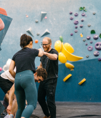 Three people at an indoor climbing gym, with one person helping another stand up. Colorful climbing holds are visible on the wall in the background. The mood appears friendly and supportive.
