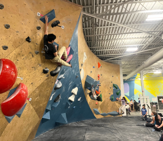Several people, including members of local youth teams, are indoor bouldering in Boston, climbing a large wooden wall with colored holds. One climber reaches high on the left as others attempt routes while onlookers watch from the padded floor under bright lights.