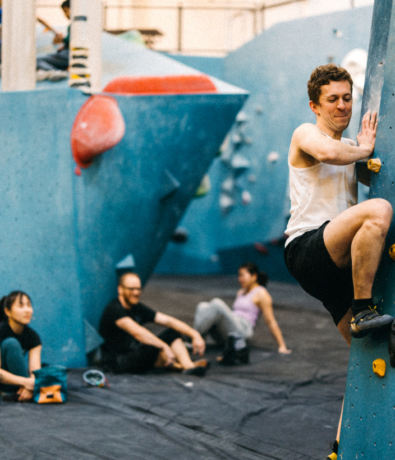 A person climbs an indoor bouldering wall while three others sit on the padded floor in the background, watching and relaxing. The scene is set in a climbing gym with blue walls and colorful holds.