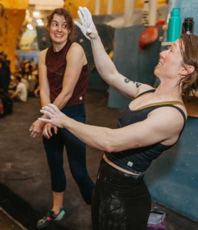 Two women in athletic wear stand in a climbing gym. One is smiling and looking at the other, who gestures with chalk-covered hands, appearing animated and expressive. Climbing walls and gear are visible in the background.