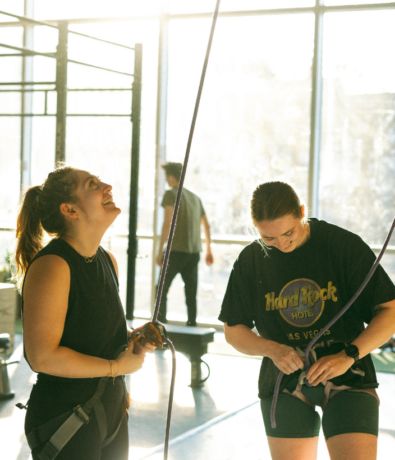 Two women in harnesses prepare for indoor climbing, smiling and standing near ropes in a sunlit gym with large windows. A person stands in the background by the window.