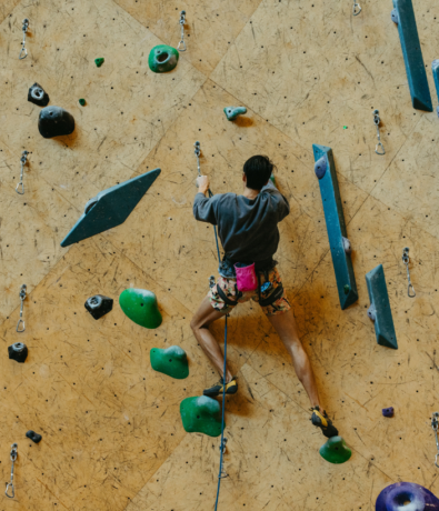 A person wearing a gray shirt, patterned shorts, and a pink chalk bag is climbing an indoor rock wall using colorful handholds and footholds, secured with a harness and rope.