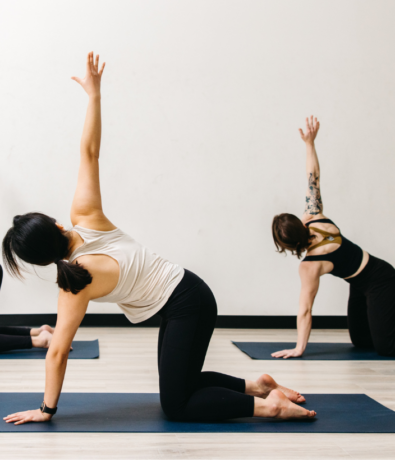 Two people practice yoga indoors on mats, kneeling and reaching one arm upward in a twist pose. They wear athletic clothing and are in a bright, minimalist studio with a light wooden floor and plain white wall.