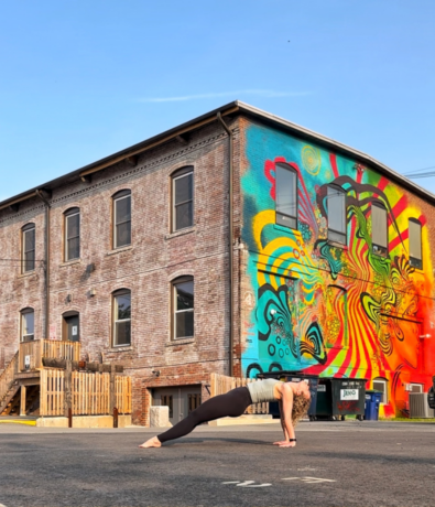 A person holds a plank pose on an empty street in front of a brick building with a vibrant, colorful mural featuring swirling abstract patterns. The sky is clear and blue.