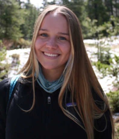 A young woman with long blonde hair smiles outdoors, wearing a black jacket and a teal scarf. Sunlight shines on her face, and a forest with trees and greenery is visible in the background.