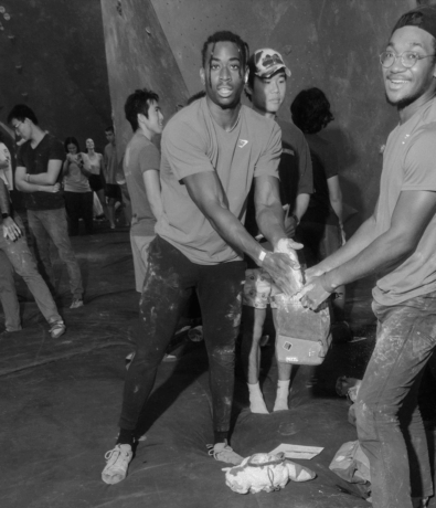 Two men smile while sharing chalk for climbing at the lively Austin Boulderfest competition in a busy indoor rock climbing gym; other climbers and onlookers stand in the background, creating a vibrant, social atmosphere.