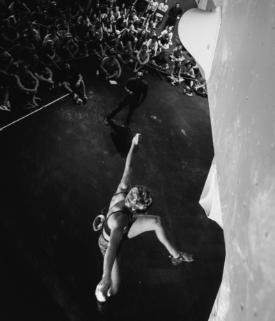 A climber reaches upward on an indoor climbing wall during the Austin Boulderfest competition, with an audience seated and watching below in the background; the image is in black and white.