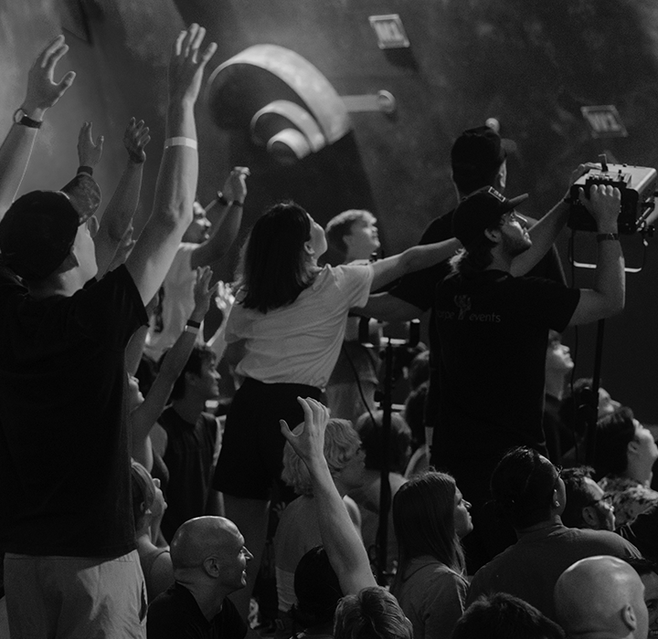 A black-and-white photo of a crowd at an indoor rock climbing gym during the Austin Boulderfest competition, with many people raising their arms and cheering, facing the climbing walls in the background.