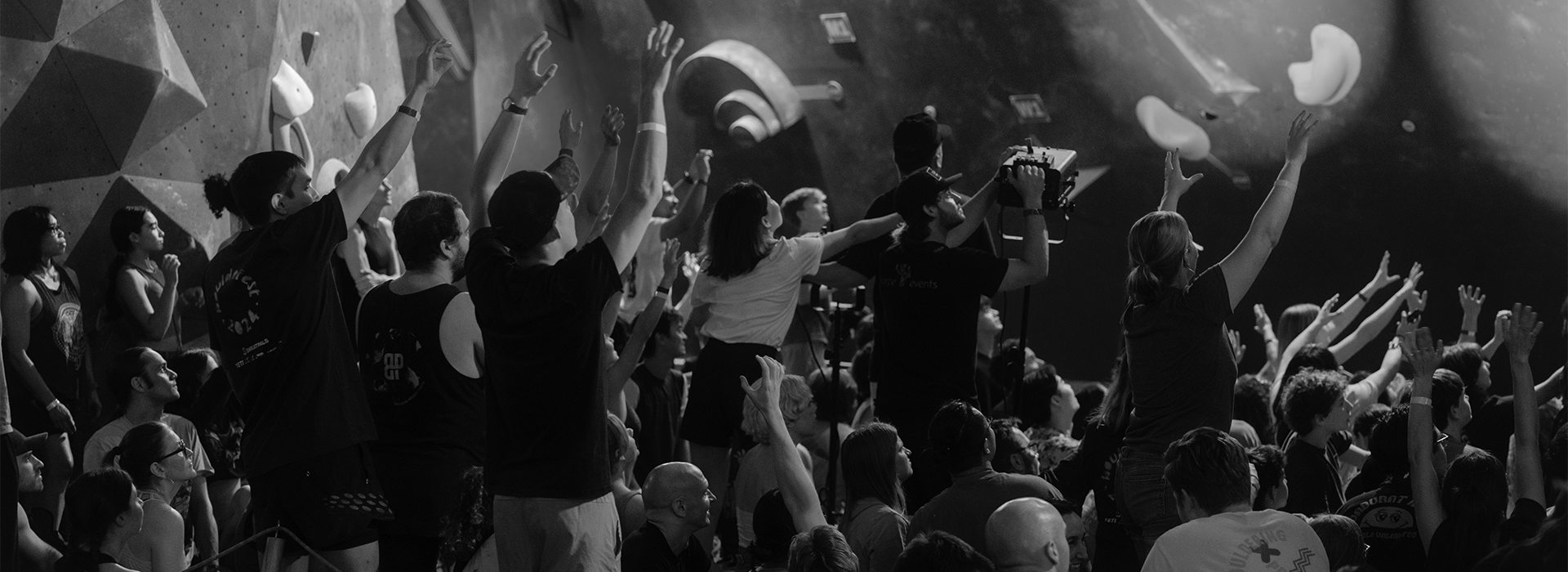 A black-and-white photo of a crowd at an indoor rock climbing gym during the Austin Boulderfest competition, with many people raising their arms and cheering, facing the climbing walls in the background.