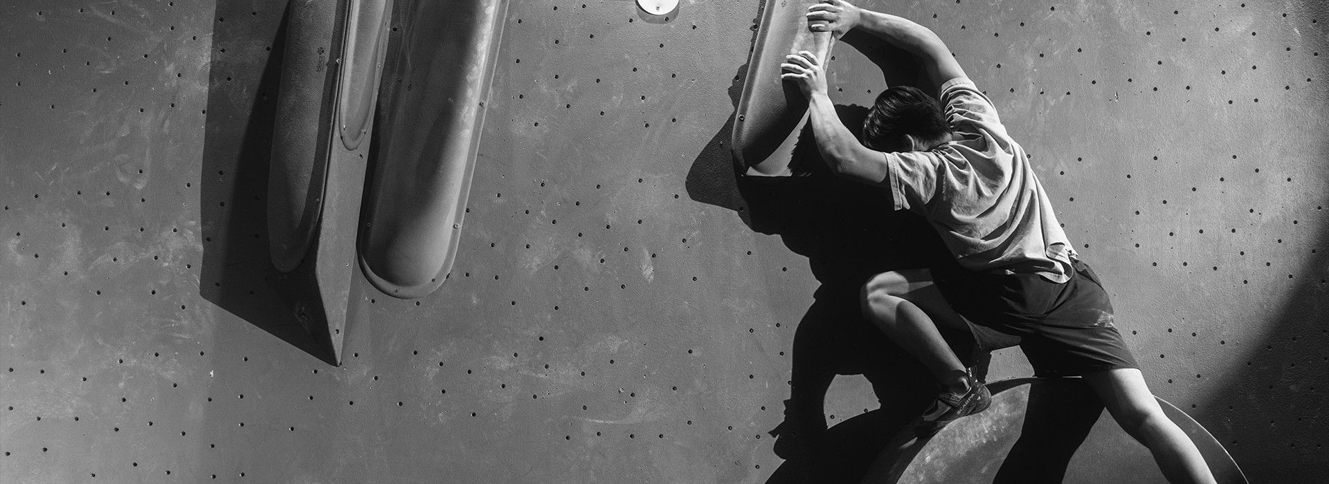 A person climbs an indoor bouldering wall at the Austin Boulderfest competition, gripping large handholds and balancing on a foothold, with dramatic shadows cast on the black and white scene.