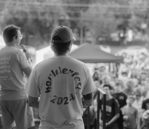 Two people stand on a stage facing a crowd at an outdoor event. One person’s shirt reads Boulderfest 2024, hinting at the exciting boulderfest competition in Austin. Tents and trees are visible in the background on a sunny day.