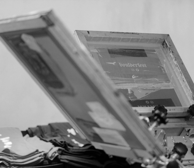 A close-up, black and white photo of a screen printing setup with a screen displaying the word boulderfest, positioned above stacked t-shirts, ready for the Austin Boulderfest competition.
