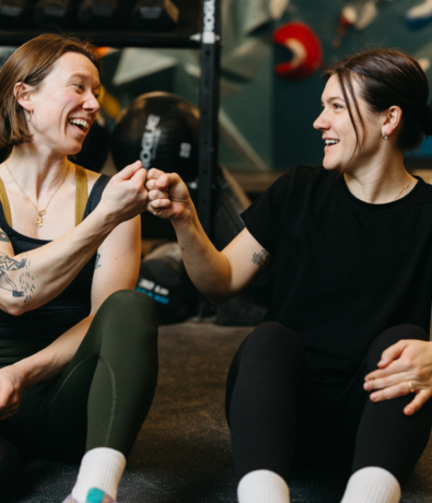 Two women sit on the Brooklyn Climbing Yoga and Fitness gym floor, smiling and giving each other a fist bump. Both wear athletic clothing, with gym equipment in the background. They appear happy and relaxed.