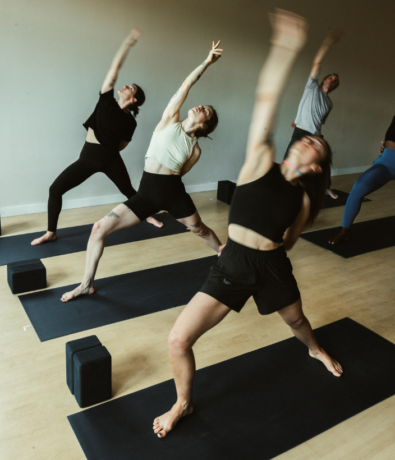 A group of people practice yoga on mats indoors at Brooklyn Climbing Yoga and Fitness, each reaching one arm upward and leaning back in a pose. Yoga blocks are placed beside them on the floor.