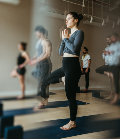 A group of people practice yoga indoors at Brooklyn Climbing Yoga and Fitness, standing on mats in tree pose with hands together at their chests. The focus is on a woman in the foreground, with others slightly blurred in the background.