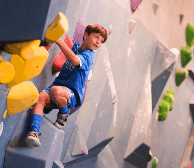 A young boy in a blue athletic outfit climbs an indoor bouldering wall during Adventure Days in Brooklyn, gripping yellow handholds and looking to the side. Colorful holds are visible around him on the gray wall.