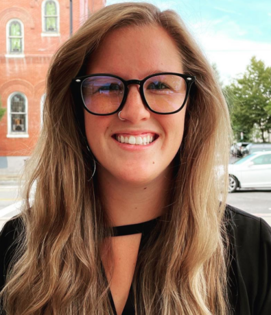 A woman with long, wavy light brown hair and black glasses smiles at the camera, sitting outdoors near a street with a brick building.