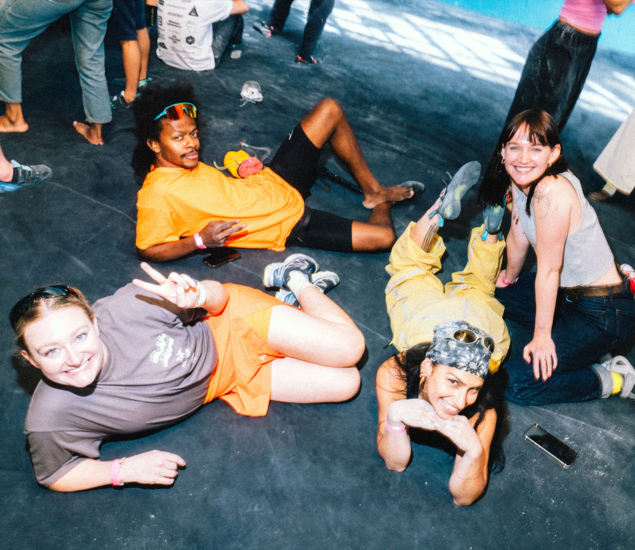 Four people sit and recline on a gym floor, smiling and posing playfully for the camera. One flashes a peace sign; another wears goggles. Climbing shoes and chalk hint at the fun vibe of Community & Events at Austin Bouldering Project.