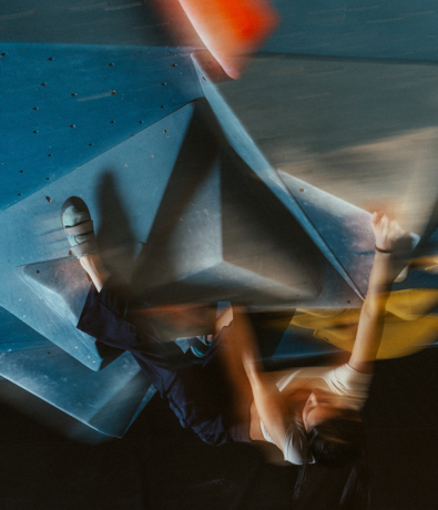 A blurred action shot of a climber wearing a white shirt and climbing shoes scaling an indoor bouldering wall with blue, gray, and yellow holds. The motion blur captures dynamic movement and intensity.