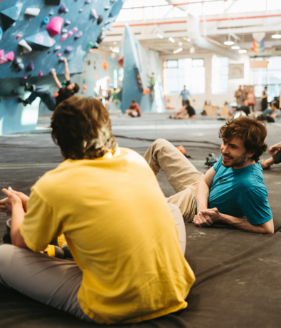 Two people sit on padded gym flooring, chatting and smiling, with one wearing a yellow shirt and the other a blue shirt. In the background, people are rock climbing on indoor climbing walls.