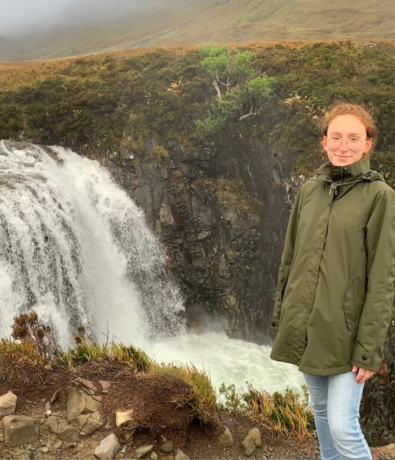 A person wearing glasses and a green raincoat stands near a rocky edge by a waterfall with lush greenery and misty hills in the background.