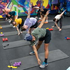 A group of people stretch on yoga mats in a colorful gym, reaching one arm up and the other to their opposite foot. Dumbbells and climbing gear are beside each mat, and a vibrant mural decorates the wall in the background.