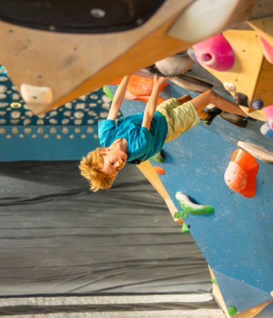 A young boy wearing a blue shirt and light shorts is climbing upside down on an indoor bouldering wall with colorful holds. Sunlight shines on him, highlighting his focus and determination.