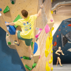 A man in a yellow shirt and green shorts enjoys climbing a colorful indoor bouldering wall, reaching for a hold. Another person stands in the background, watching or waiting their turn.