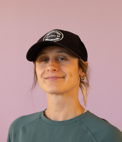A person with long hair tied back, wearing a black Hurley cap, gold hoop earrings, and a green top, smiles softly against a light purple background at an Austin Climbing Gym.