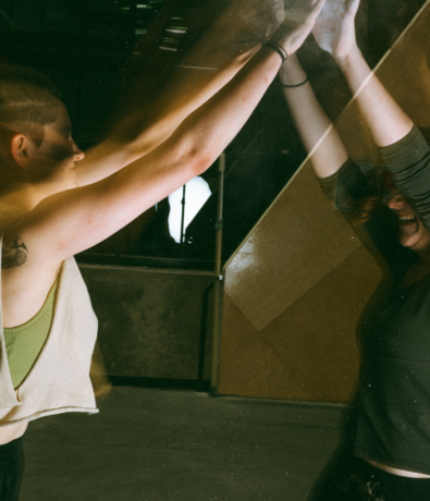 Two people stand facing each other, separated by a pane of glass. As they both raise their hands to touch the glass, it creates a mirrored effect with their reflections—almost as if theyre climbing invisible boundaries in the warm, soft light.