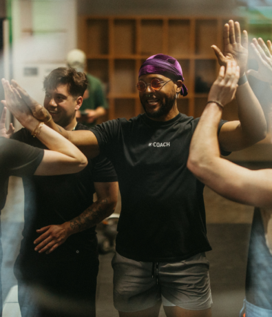 A group of people smiling and giving each other high fives in a climbing gym. One person in the center wears a purple headscarf, glasses, and a shirt that says COACH. They appear happy and energized.