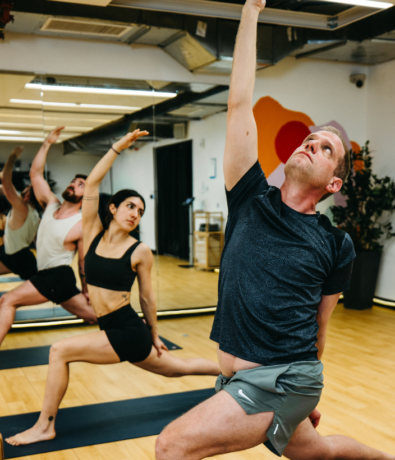 A group of people practice yoga in a studio, performing a lunge pose with one arm extended upward, as if reaching toward a climbing hold. The room features wooden floors, mirrors, and plants in the background.