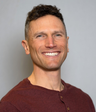 A smiling man with short brown hair wearing a maroon shirt stands against a plain light gray background, ready to offer top-notch personal training in SLC.