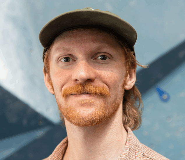 A person with light skin, red hair, and a mustache is wearing a green cap and a checked shirt, standing in front of a blurred blue and gray background—perfectly capturing the approachable vibe of Personal Climbing Coaching Austin.