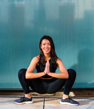 A woman in athletic wear is squatting with her legs wide apart and hands pressed together in a prayer position. She is smiling and outdoors, with a teal wall in the background.