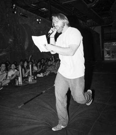 A man with shoulder-length hair, beard, and glasses holds a microphone and paper while speaking to a seated crowd indoors. He stands on one leg, mid-step, in a casual pose on the padded floor of an Austin climbing gym.