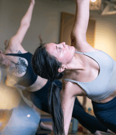 Two people practicing yoga indoors at an Austin climbing gym; the person in front, wearing a light sports bra and dark leggings, stretches upward in a side angle pose, focusing intently. Another person is visible in the background.