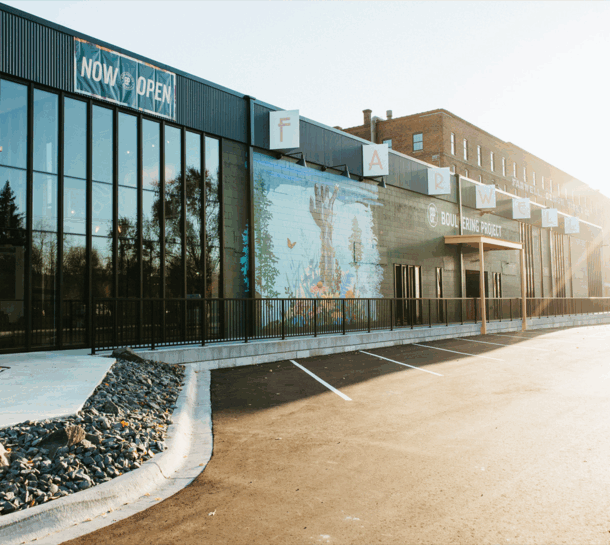 A modern building with large glass windows, a mural of hands and plants, a NOW OPEN sign, and an empty parking lot in front. The Boulder Food Project sits next to the St. Paul Bouldering Project Climbing Gym.