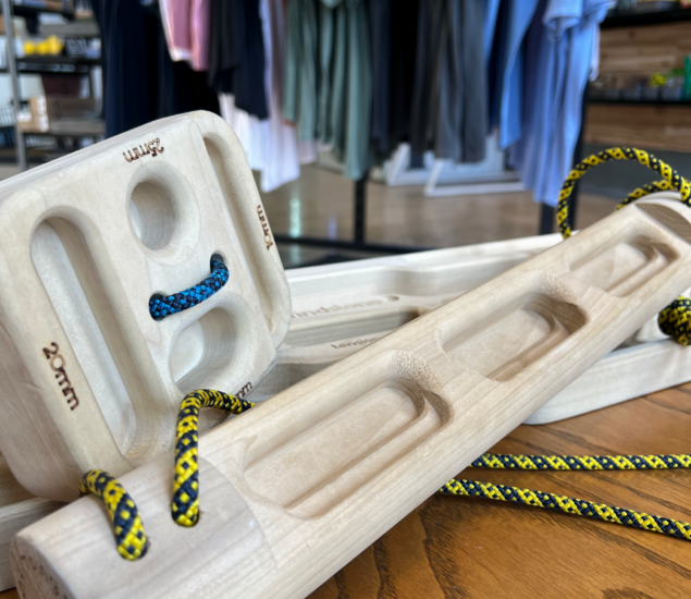 Close-up of MBP wooden hangboards with ropes, used for climbing training, displayed on a wooden table. Shirts hang blurred in the background inside a retail store.