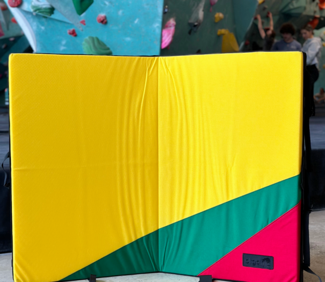 A colorful climbing crash pad with yellow, green, and red sections stands open in front of an indoor climbing wall at Retail at MBP. Climbers and holds are visible in the background.