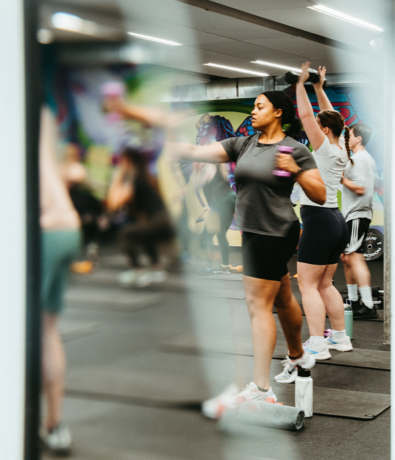 A group of people exercise in a fitness class, holding small dumbbells and stretching their arms upward and outward. The scene is energetic, and a colorful mural is visible on the wall behind them.