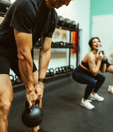 Two people exercising in a gym; the man in the foreground is holding a kettlebell, while the woman in the background is performing a squat with a kettlebell. Both are wearing workout clothes.