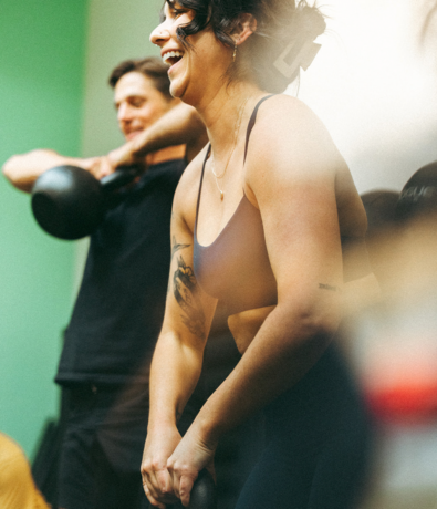 Two people smiling and lifting kettlebells in a brightly lit gym, appearing energetic and engaged in a workout. The focus is on the woman in the foreground with short dark hair and a tattoo on her arm.