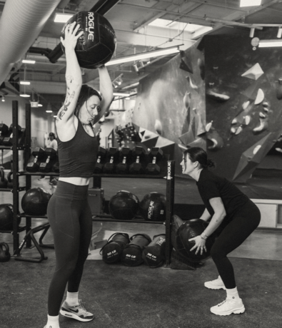 Two women exercise in a Design District gym. One stands, lifting a medicine ball overhead, while the other squats holding a ball. Climbing walls and weights appear in the black and white background.